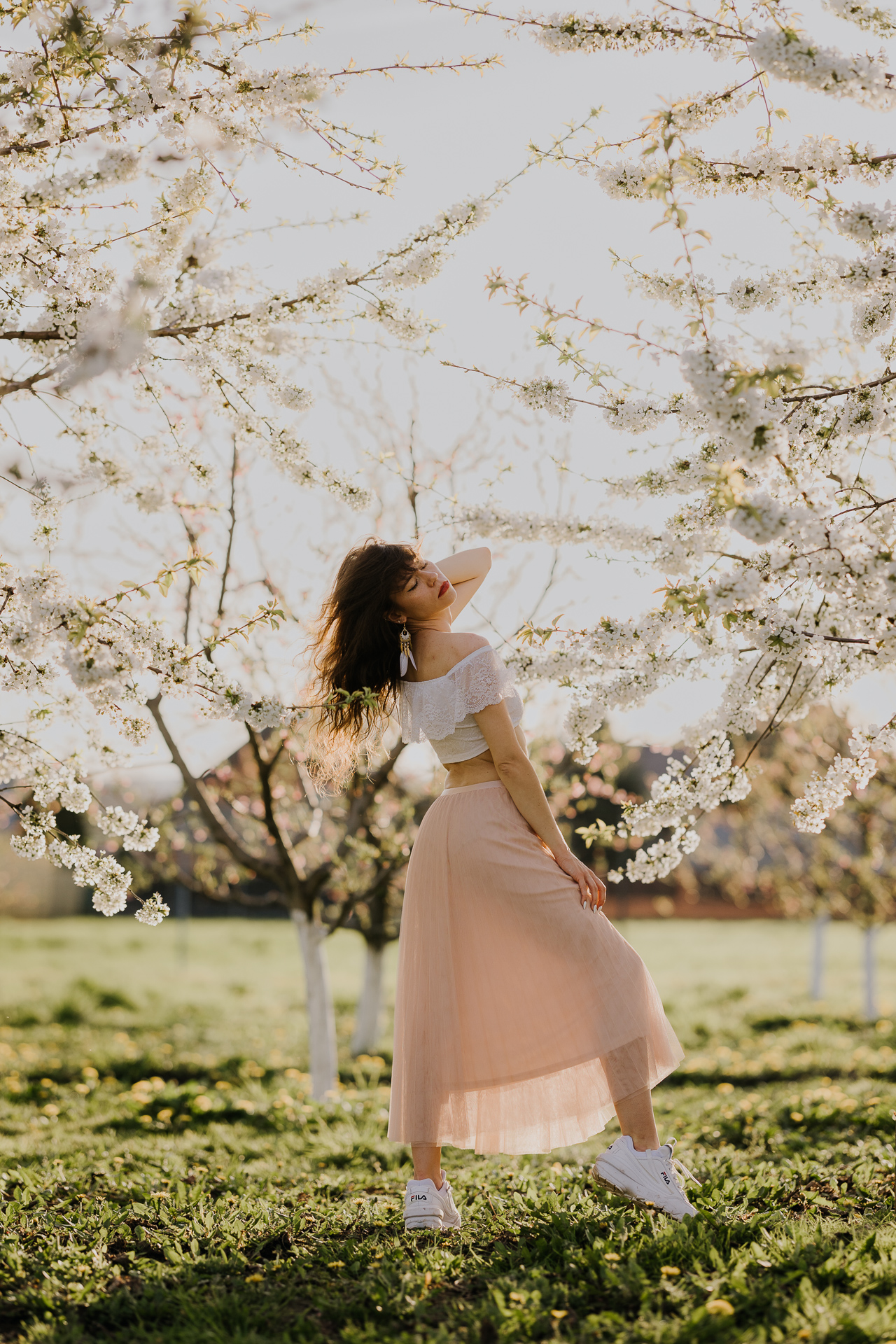 Kobieta w białej bluzce i różowej spódnicy na tle kwitnących drzew owocowych, sesja fotograficzna wiosenna w sadzie pod Krakowem