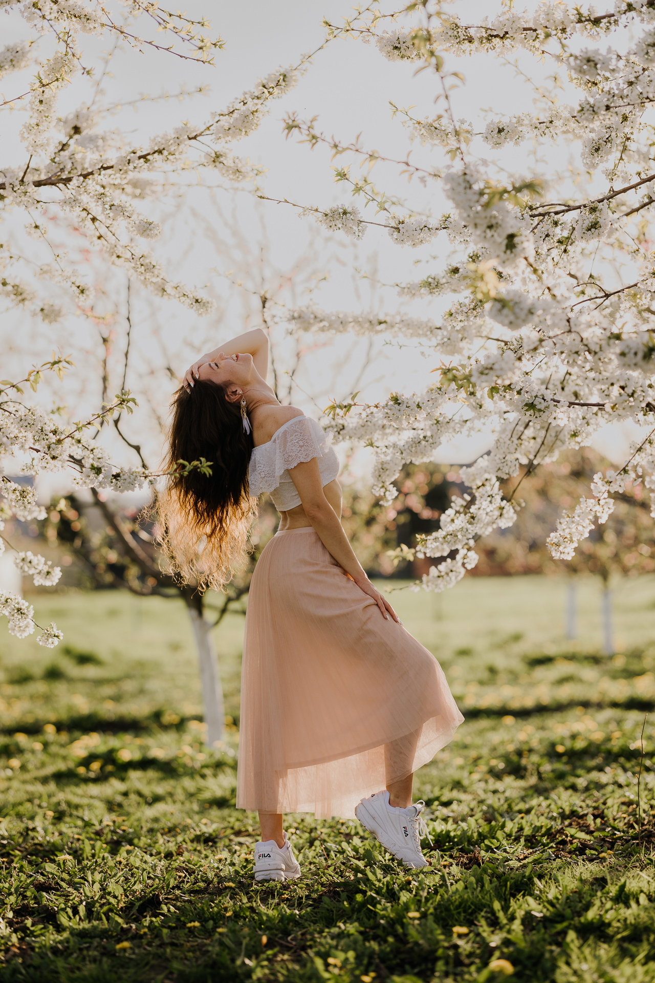 Uśmiechnięta Kobieta w białej bluzce i różowej spódnicy na tle kwitnących drzew owocowych, sesja fotograficzna wiosenna w sadzie pod Krakowem