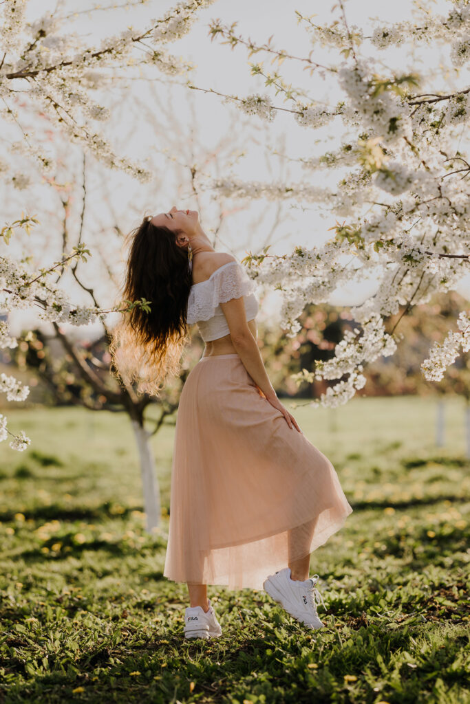 Uśmiechnięta Kobieta w białej bluzce i różowej spódnicy na tle kwitnących drzew owocowych, sesja fotograficzna wiosenna w sadzie pod Krakowem