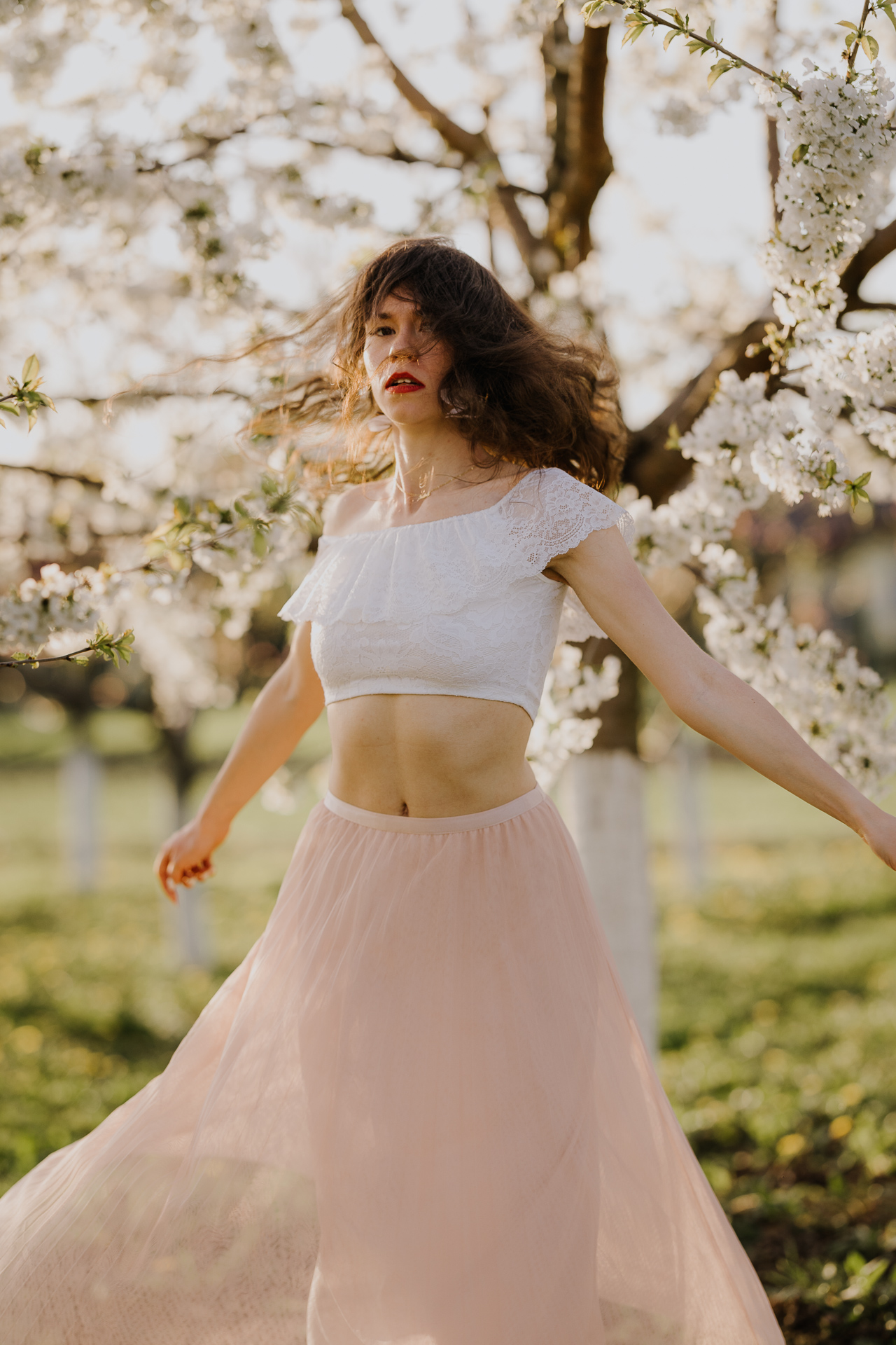 Tańcząca Kobieta w białej bluzce i różowej spódnicy na tle kwitnących drzew owocowych, sesja fotograficzna wiosenna w sadzie pod Krakowem