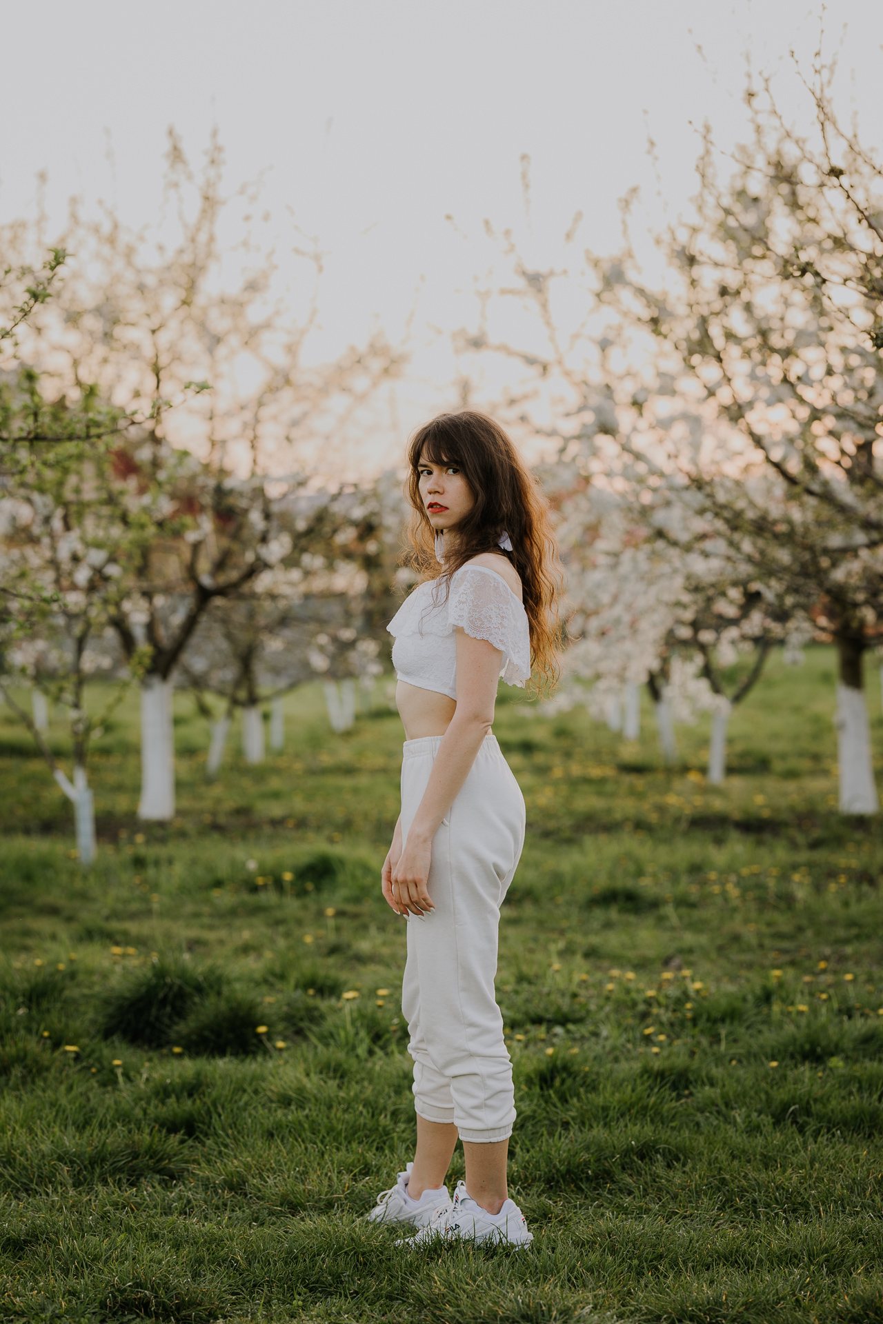 Kobieta w białej bluzce i spodniach na tle kwitnących drzew owocowych, sesja fotograficzna wiosenna w sadzie pod Krakowem