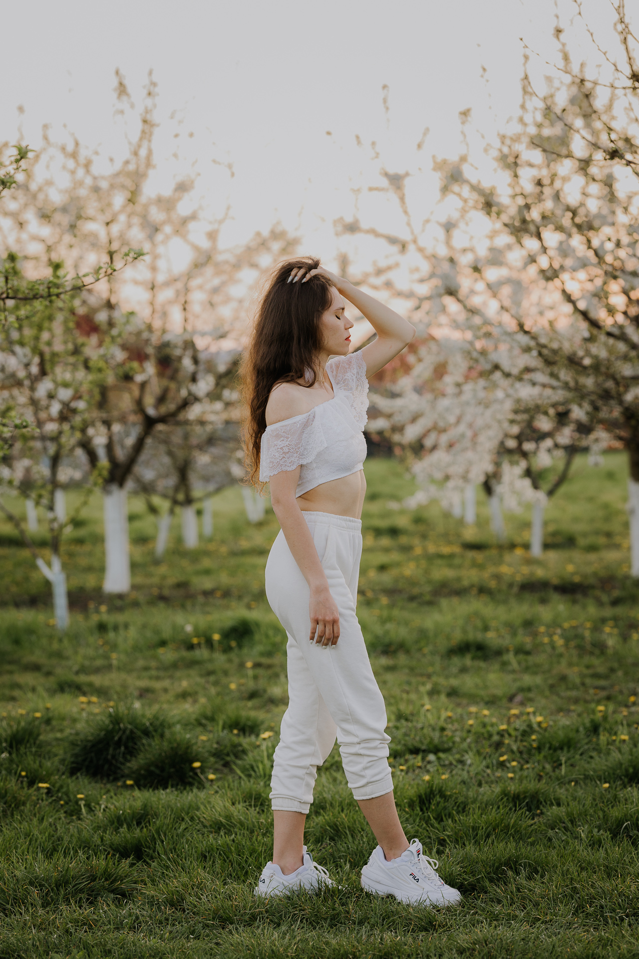 Kobieta w białej bluzce i spodniach na tle kwitnących drzew owocowych, sesja fotograficzna wiosenna w sadzie pod Krakowem