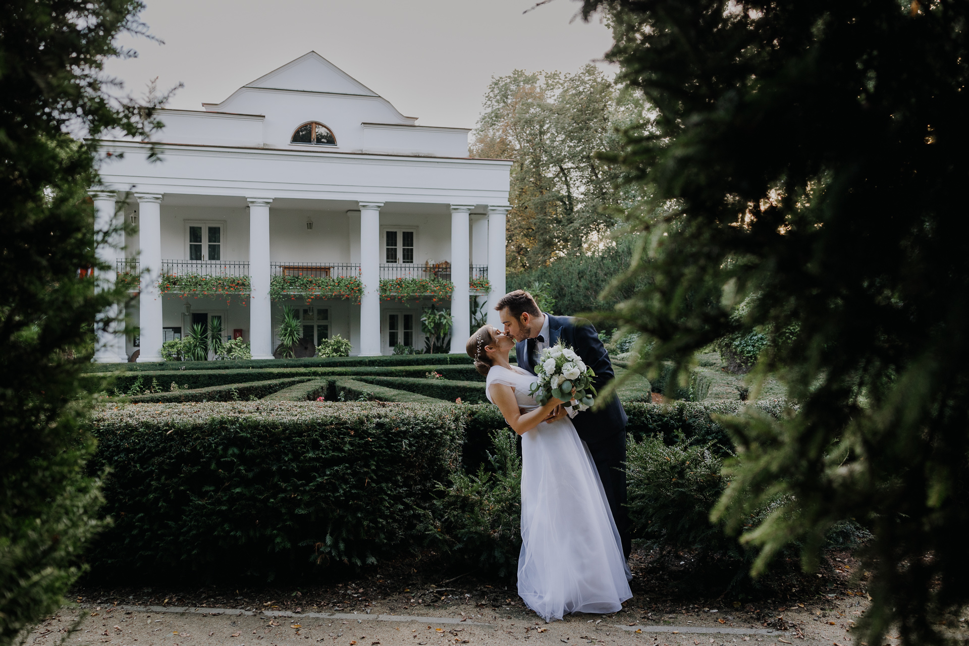 Para młoda całuje się na tle budynku w Arboretum Bolestraszyce, romantyczne ujęcie wśród roślin, fotografia ślubna Podkarpacie