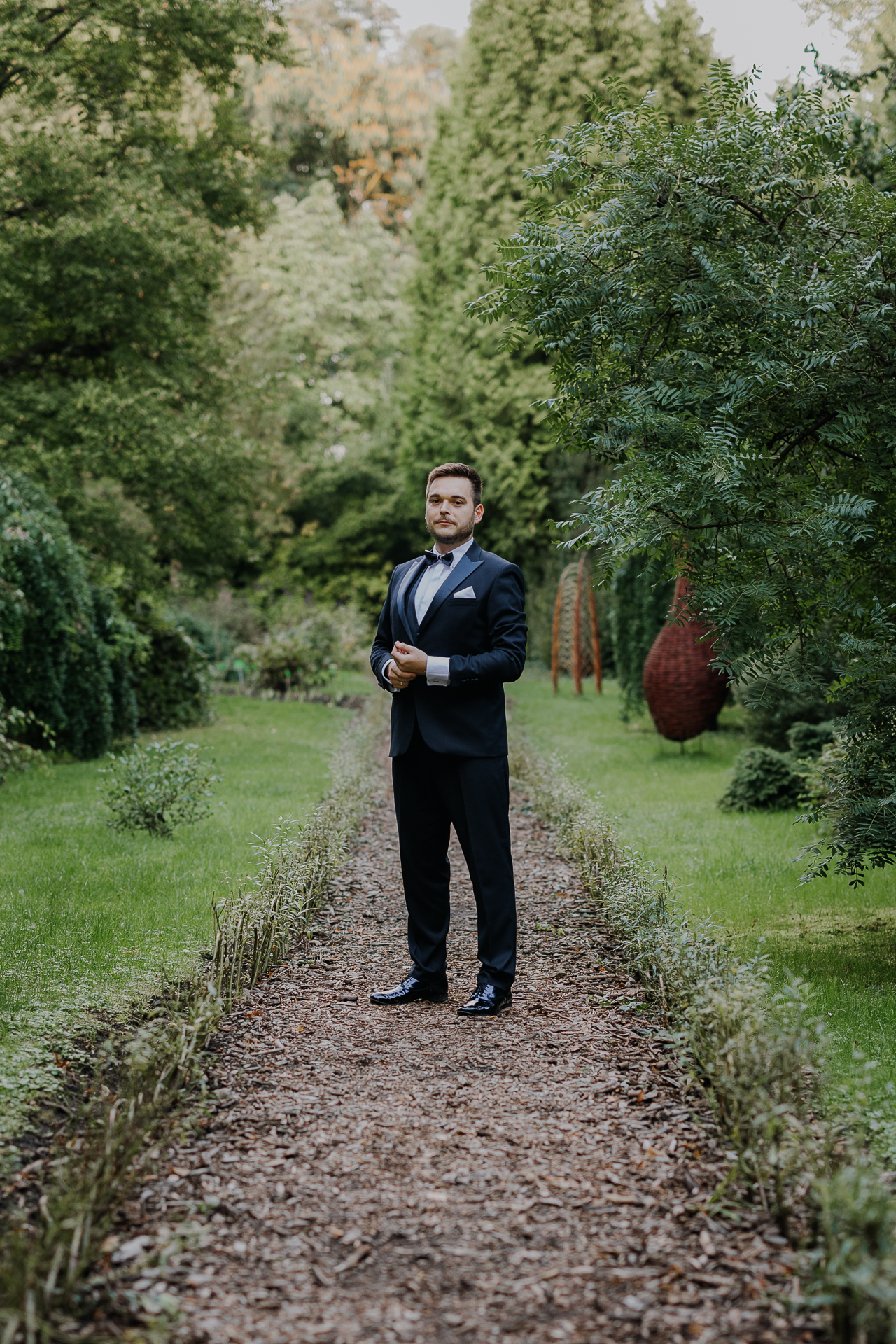 Portret pana młodego w eleganckim garniturze na tle bujnej roślinności arboretum Bolestraszyce, męska fotografia ślubna