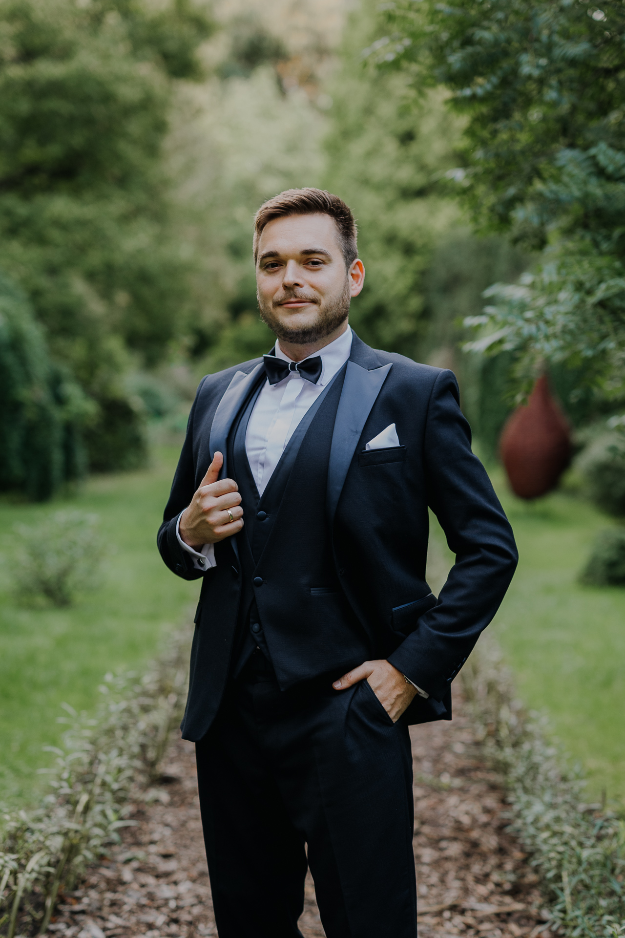 Portret pana młodego w eleganckim garniturze na tle bujnej roślinności arboretum Bolestraszyce, męska fotografia ślubna