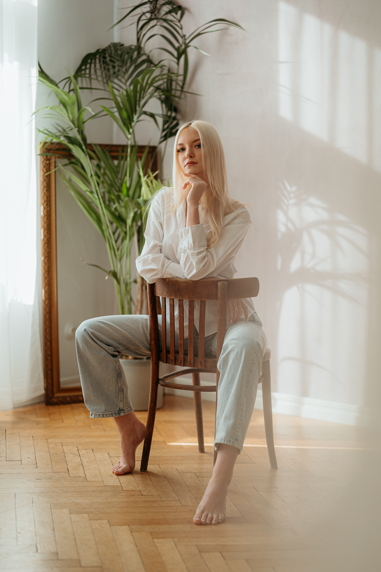 Portret kobiety siedzącej na krześle z ręką pod brodą, miękkie światło wpadające przez okno tworzy wiosenną atmosferę sesji