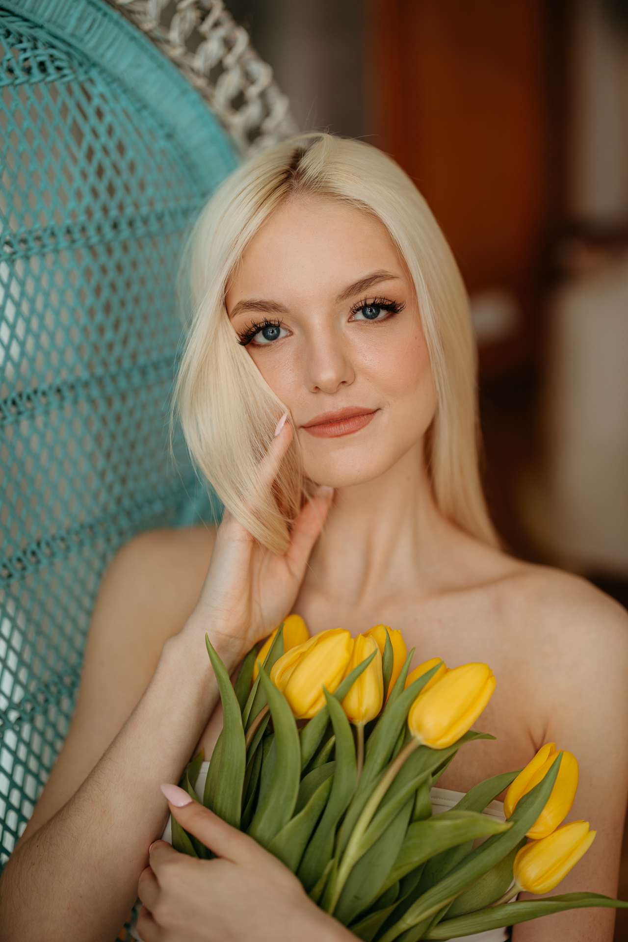 Portret kobiety siedzącej na krześle z żółtymi tulipanami w dłoniach, miękkie światło wpadające przez okno tworzy wiosenną atmosferę sesji