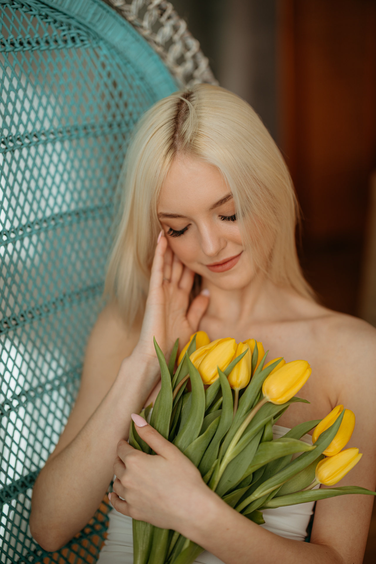 Portret kobiety siedzącej na krześle z żółtymi tulipanami w dłoniach, miękkie światło wpadające przez okno tworzy wiosenną atmosferę sesji