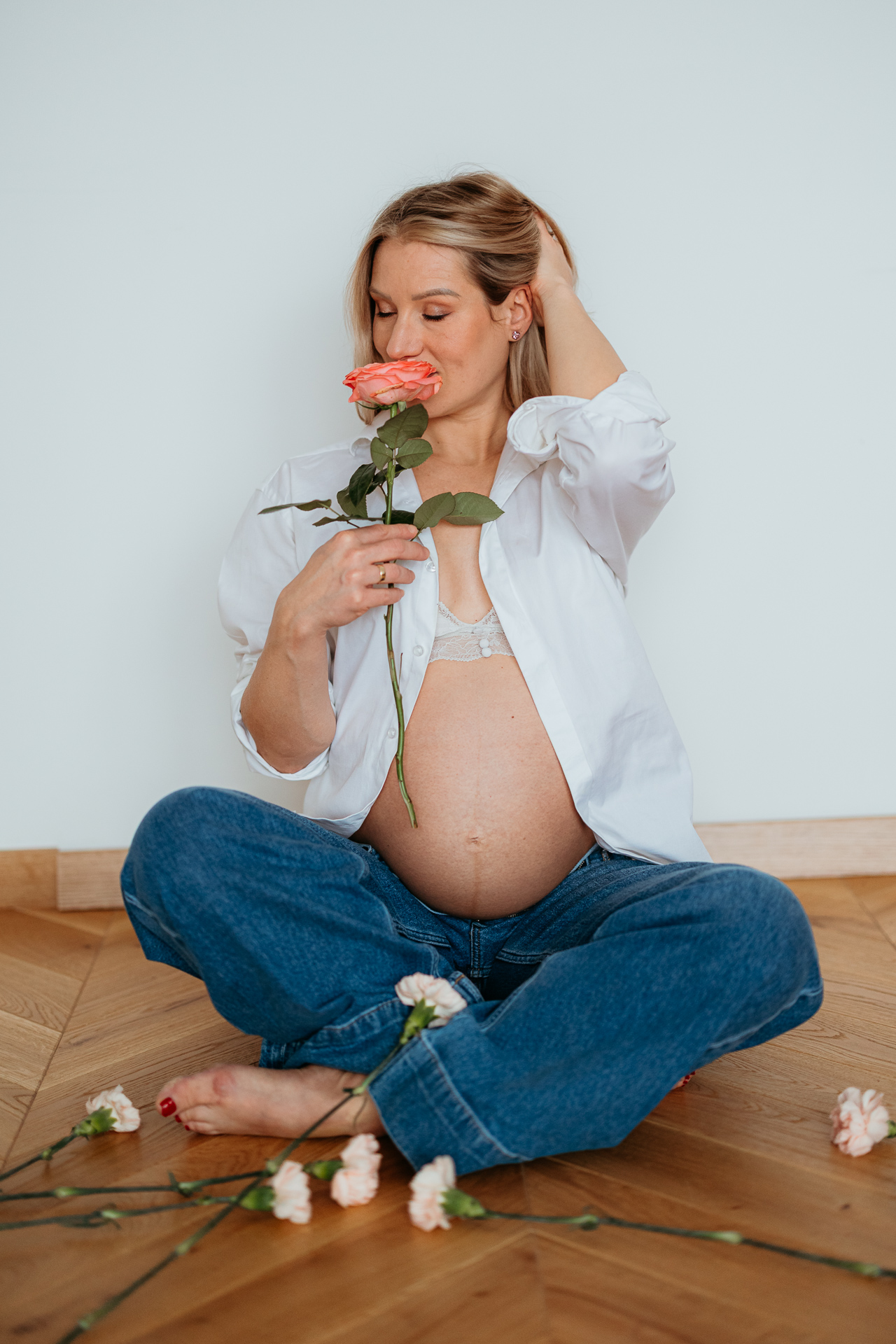 Uśmiechnięta kobieta z odsłoniętym brzuszkiem ciążowym siedzi na podłodze i trzyma różę przy buzi w domu w Krakowie