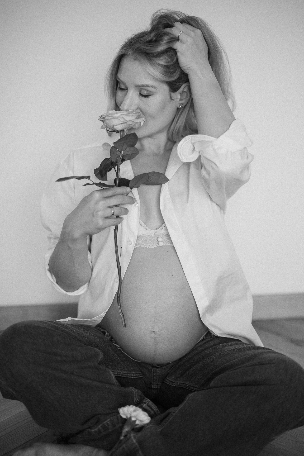 Uśmiechnięta kobieta z odsłoniętym brzuszkiem ciążowym siedzi na podłodze i trzyma różę przy buzi w domu w Krakowie