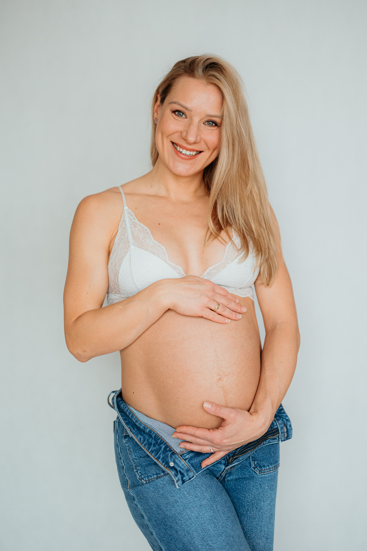 Uśmiechnięta kobieta w białym biustonoszu z odsłoniętym brzuszkiem, trzyma się za brzuszek ciążowy w Krakowie