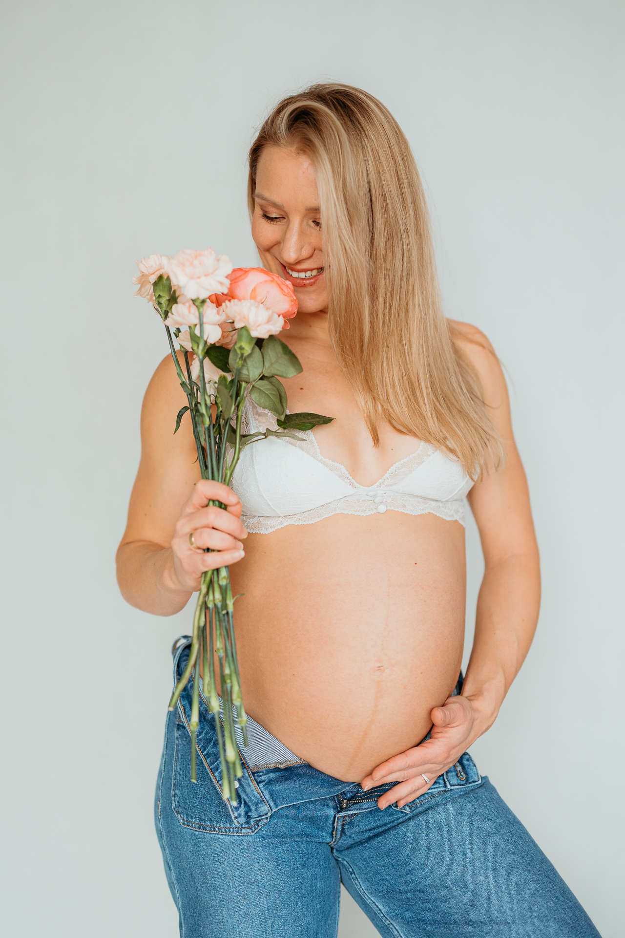 kobieta w białym biustonoszu z odsłoniętym brzuszkiem i różami w ręce trzyma się za brzuszek ciążowy w Krakowie