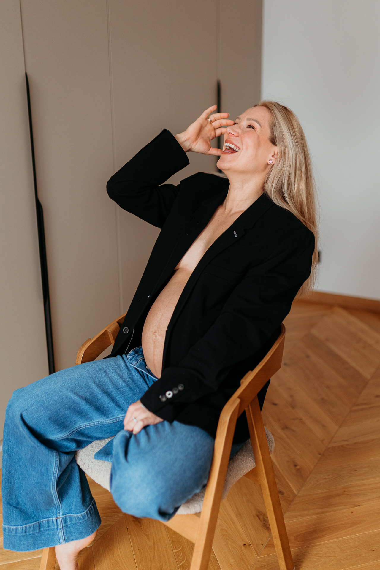 Roześmiana kobieta siedzi w czarnej marynarce i jeansach z odsłoniętym brzuszkiem w domu w Krakowie