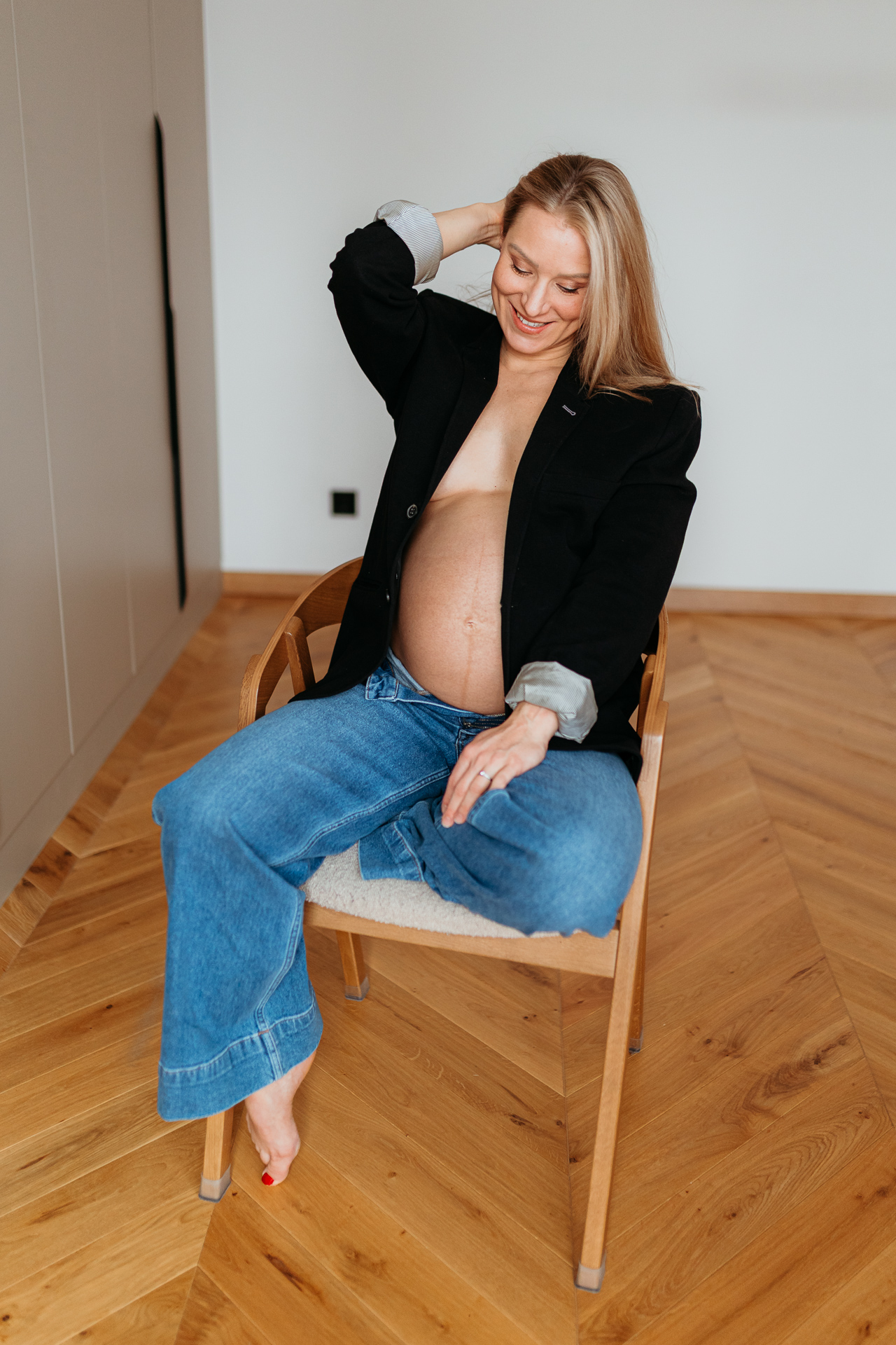 Roześmiana kobieta siedzi w czarnej marynarce i jeansach z odsłoniętym brzuszkiem w domu w Krakowie