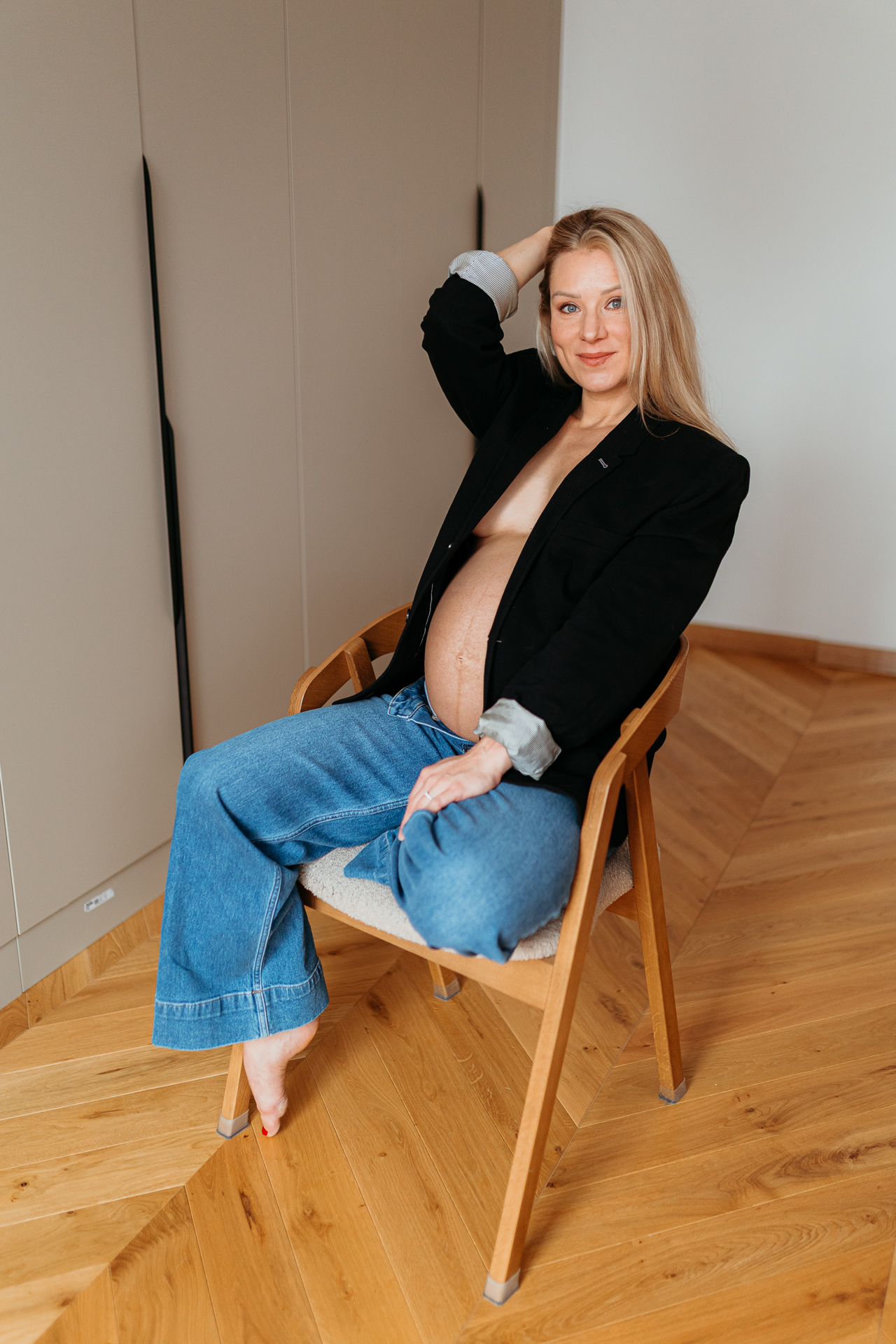 Roześmiana kobieta siedzi w czarnej marynarce i jeansach z odsłoniętym brzuszkiem w domu w Krakowie