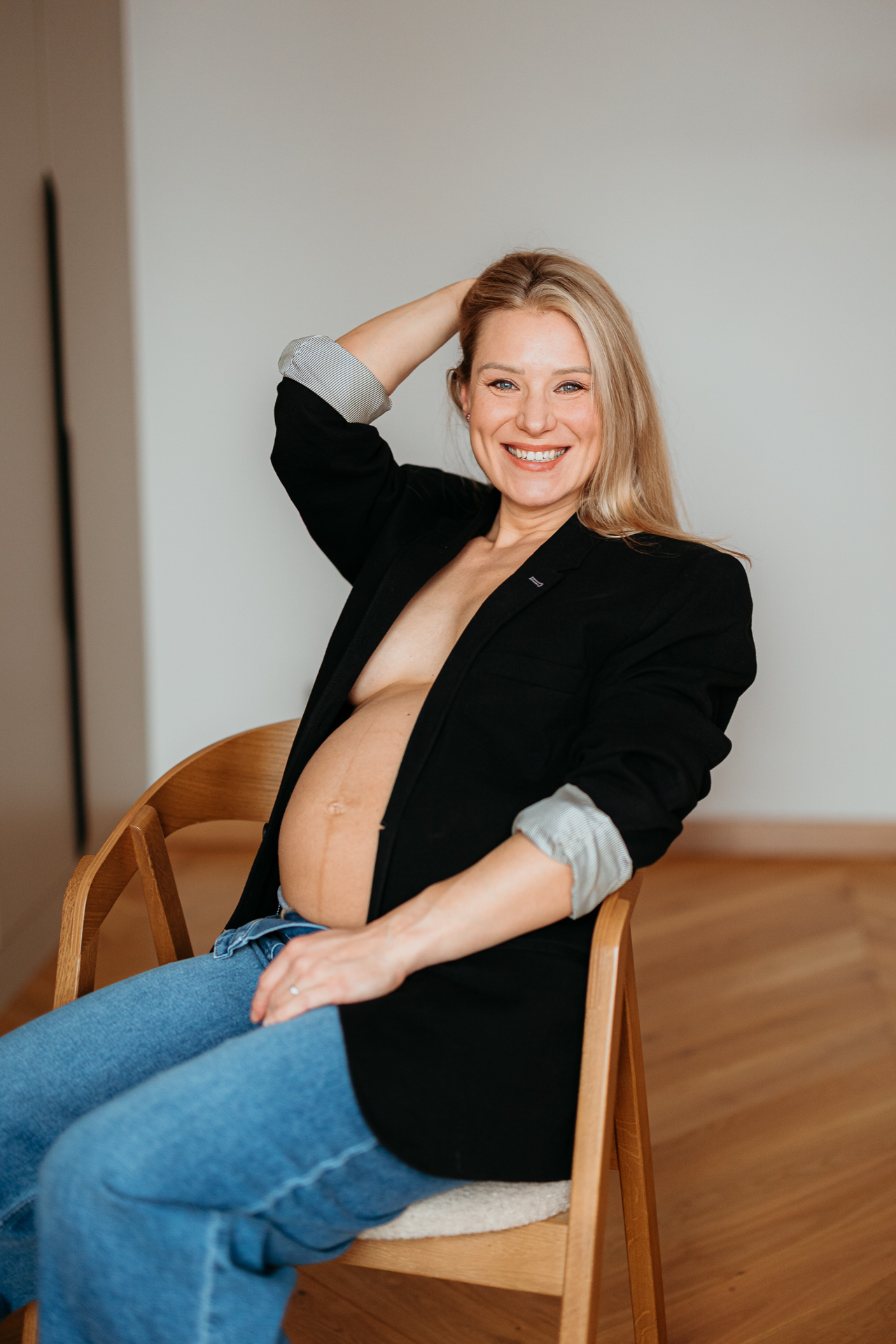 Roześmiana kobieta siedzi w czarnej marynarce i jeansach z odsłoniętym brzuszkiem w domu w Krakowie