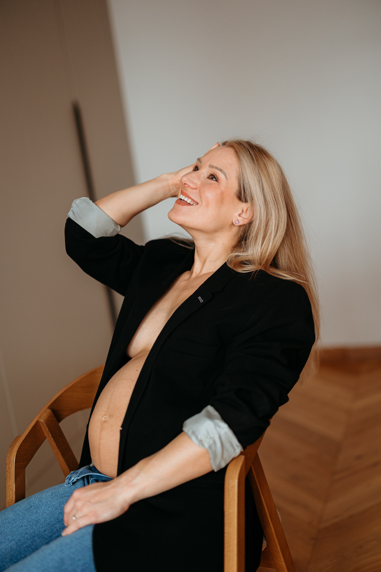 Roześmiana kobieta siedzi w czarnej marynarce i jeansach z odsłoniętym brzuszkiem w domu w Krakowie