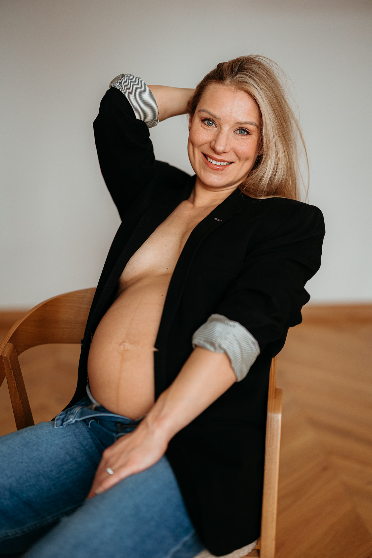 Roześmiana kobieta siedzi na krześle w czarnej marynarce i jeansach z odsłoniętym brzuszkiem w domu w Krakowie