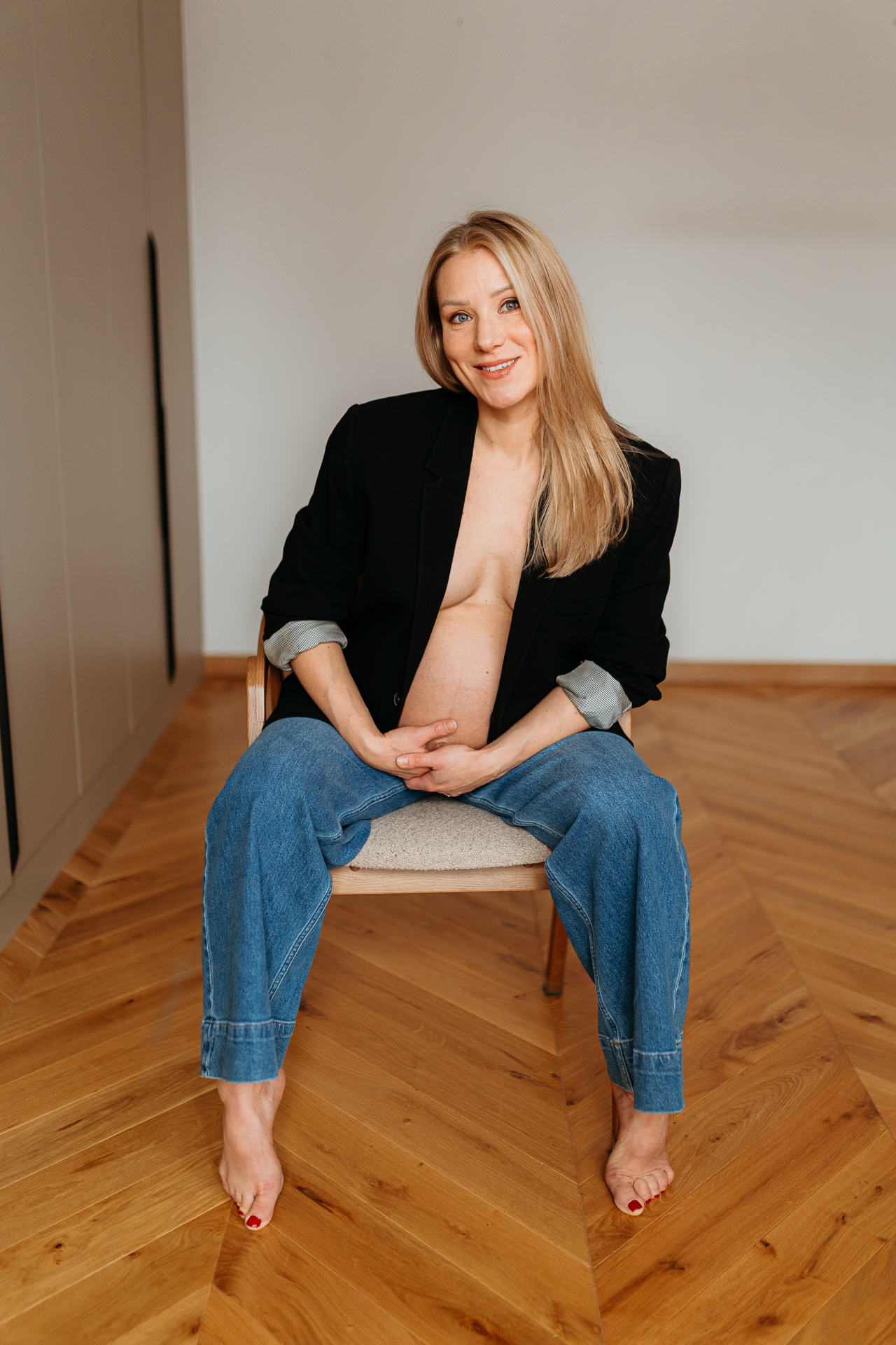 Uśmiechnięta kobieta siedzi w czarnej marynarce i jeansach z odsłoniętym brzuszkiem w domu w Krakowie